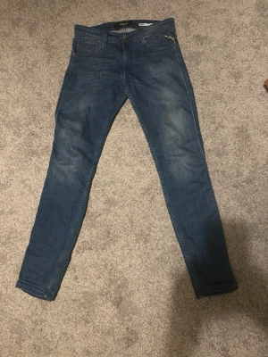 Replay Anbass jeans blå - Säljer ett par blå Replay Anbass jeans i slim fit och snygga slitningar. Jeansen har mellanblå tvätt, dragkedjegylf och detaljerad bakficka med Replay-logga. Perfekta för dig som gillar en smalare passform och stilren look. nypris runt 1100kr