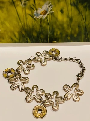 Blommigt armband med myntdetaljer - Unikt armband i silverfärgad metall med stora blommor och hängande guldfärgade mynt med asiatiska tecken. Länkkedja och karbinlås gör det enkelt att justera längden. Perfekt för dig som gillar boho och statement-smycken.