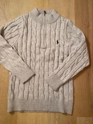 Beige kabelstickad tröja Polo Ralph Lauren - Säljer en beige kabelstickad tröja från Polo Ralph Lauren med halv dragkedja och ribbade muddar. Tröjan har det klassiska broderade logotypen på bröstet och är gjord i mjukt material. Perfekt för dig som gillar stilren och tidlös design.