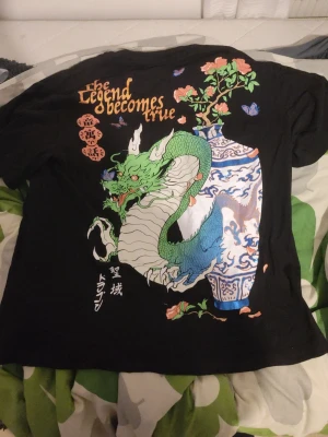 Svart t-shirt med draktryck - Cool svart t-shirt med stort färgglatt tryck på ryggen som visar en grön drake, blåvit vas och blommor. Texten 'The legend becomes true' och japanska tecken ger en unik vibe. Perfekt för dig som gillar streetstyle och asiatiska influenser.
