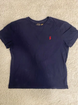 Mörkblå t-shirt från Polo Ralph Lauren - Mörkblå t-shirt från Polo Ralph Lauren med klassisk röd broderad logga på bröstet. T-shirten har rund halsringning och är tillverkad i mjuk bomull. Perfekt för en stilren och enkel look.