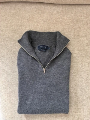 Grå halvzip i merinoull från Riley - Snygg grå stickad tröja från Riley i mjuk merinoull. Tröjan har halv dragkedja och hög krage, perfekt för lager på lager. Enkel och stilren design som passar till det mesta. Materialet är 100% merinoull vilket gör den både varm och andas bra.