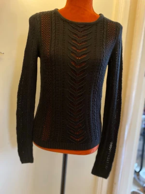 Svart stickad tröja med hålmönster XS/S - Snygg svart stickad tröja med lång ärm och dekorativt hålmönster framtill och på ärmarna. Tröjan har rund halsringning och är figurnära i modellen. Perfekt för lager-på-lager och ger en trendig look.