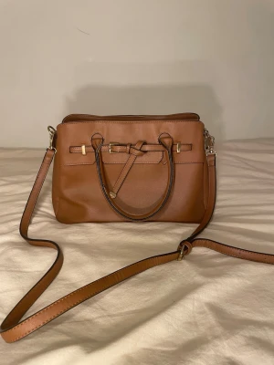 Brun axelväska med gulddetaljer - Snygg brun axelväska i slätt konstläder med justerbar axelrem och två handtag. Väskan har guldfärgade metalldetaljer och dekorativ knut framtill. Perfekt storlek för det viktigaste och stängs med dragkedja upptill.