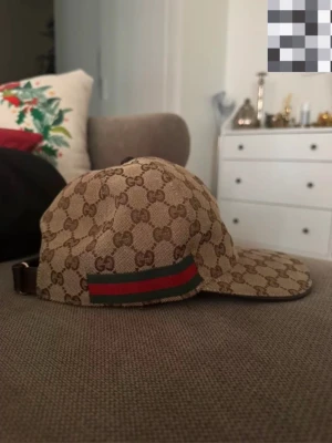 Gucci beige keps med GG-mönster - Pris är diskuterbart.!  Säljer en beige keps från Gucci med det klassiska GG-monogrammönstret över hela kepsen. Kepsen har en grön och röd rand på sidan och justerbart spänne bak i brunt läder. Perfekt för dig som vill ha en stilren och lyxig accessoar. 