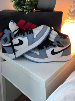 Nike Air Jordan 1 Mid University Blue - S soppar ett par Nike Air Jordan 1 Mid sneakers i grått, blått och vitt med svarta detaljer. Klassisk Swoosh-logga på sidan, högt skaft och vadderad plös. Ovandel i läder och textil, yttersula i gummi. Perfekta för dig som gillar streetwear och ikonisk sneakerstil. Om priset är för mycket ge ett förslag.