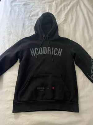Svart hoodie från Hoodrich - Snygg svart hoodie från Hoodrich med stor logga på bröstet och tryck på ärmen. Klassisk känguruficka framtill och justerbar huva med snören. Perfekt för en streetwear-look och riktigt skön att bära. Materialet känns mjukt och bekvämt.