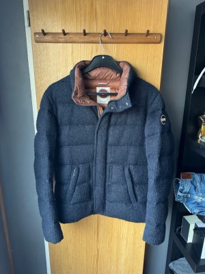 Mörkblå pufferjacka från Colmar - Säljer en mörkblå pufferjacka från Colmar med manschetter yta och brunt innerfoder. Jackan har hög krage, två framfickor och Colmar -logga på ärmen. Perfekt för kalla vinterdagar och riktigt snygg till jeans. Den har lite tecken på användning såsom hålen där man knäpper jackan