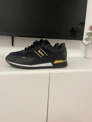 Svarta BOSS sneakers med gulddetaljer - Stilrena svarta sneakers från BOSS med snygga gulddetaljer och logga på sidan. Skorna har lågt skaft, svarta snören och en vit sula med svarta och guldiga inslag. Materialmix av textil och syntet ger en modern look som passar till många outfits.