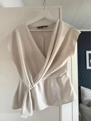 Topp från Zara - Snygg beige omlottopp från H&M med knyt i sidan och v-ringad hals. Toppen har korta ärmar och en mjuk, lätt glansig yta som ger en elegant känsla. Perfekt att styla med jeans eller kjol för en trendig look.