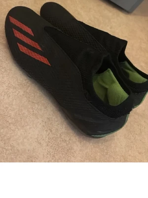 Svarta Adidas fotbollsskor utan snörning - Den har bara ett litet hål framme. Säljer ett par svarta fotbollsskor från Adidas med röda detaljer och klassiska tre ränder på sidan. Skorna har en strumpliknande passform utan snörning och insida i grönt. Perfekta för dig som vill ha bra grepp och stil på planen.