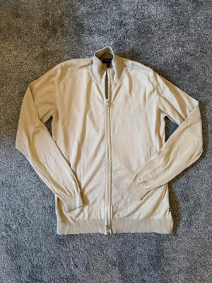 Beige ziptröja  - Stilren beige ziptröja med hög krage och långa ärmar. Tröjan har ribbade muddar vid ärmslut och nederkant samt dragkedja framtill. Perfekt för lager på lager och enkel att matcha med olika outfits. Inga defekter finns på plagget.