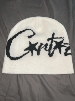 Cortez mössa - Säljer en vit mössa från Corteiz med stor svart logga i graffiti-stil på framsidan. Mössan är ribbstickad och har en mjuk, stretchig passform som sitter skönt på huvudet. Perfekt för dig som gillar streetwear och vill sticka ut.