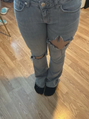 Grå jeans med hål - Säljer ett par grå jeans med hål på båda benen. Jeansen har låg- mallan längd på  midja och utsvängda ben. 
