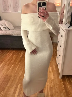 Vit offshoulder stickad långklänning - Superfin vit stickad långklänning med offshoulder-design och långa ärmar. Klänningen har en bred ribbad kant upptill som ger en snygg och trendig look. Perfekt för dig som vill ha en elegant och mysig outfit.