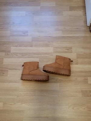 Bruna fårskinnskängor med platåsula - Säljer ett par bruna kängor i mocka-look med vit fluffig insida och rejäl platåsula. Kängorna har en klassisk, enkel design och är perfekta för att hålla värmen under kalla dagar. Passar dig som gillar en avslappnad och trendig stil.