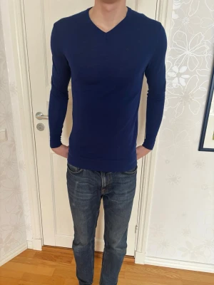 Mörkblå långärmad tröja, J.Lindeberg - Snygg mörkblå långärmad tröja med V-ringning. Tröjan har en slim fit passform och är tillverkad i ett mjukt, stretchigt material som sitter skönt mot kroppen. Perfekt att matcha med jeans för en clean look.