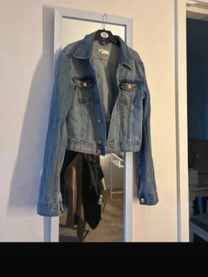  ljusblå jeansjacka - Snygg ljusblå jeansjacka. Jackan har två bröstfickor med lock och knappar framtill i kopparfärg. Perfekt att slänga över en hoodie eller t-shirt för en cool look.