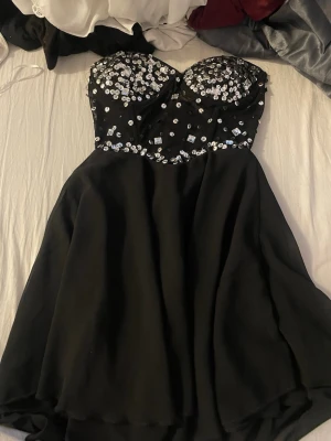 Svart kort klänning med paljetter - Säljer en svart kort klänning med strapless design och glittriga paljetter på överdelen. Kjolen är utsvängd och tyget känns lätt och följsamt. Perfekt för dig som vill sticka ut med en elegant look och massa bling.