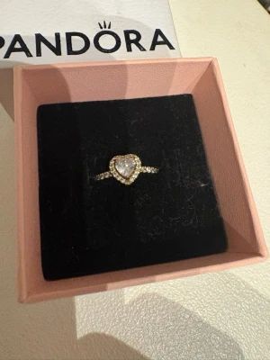 Pandora promise ring med hjärtformad sten - Snygg ring från Pandora i guld med en stor hjärtformad klar sten i mitten, omgiven av små gnistrande stenar runt om. Ringen har en elegant och romantisk design som verkligen sticker ut. Perfekt för dig som gillar smycken med det lilla extra. Den är köpt för 1100 kr och är i bra skick, perfekt som promise ring eller som en vardaglig ring. Den är i storlek 54 eller 17. 