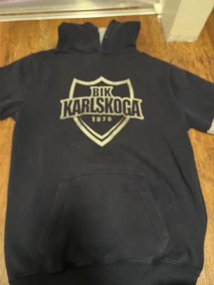 BIK Karlskoga hoodie marinblå - Marinblå hoodie från Just Hoods med stort BIK Karlskoga-tryck på bröstet. Klassisk känguruficka framtill och mjukt, borstat innerfoder. Huva utan dragsko och ribbade muddar vid ärmar och nederkant. Perfekt för dig som gillar sportig stil.