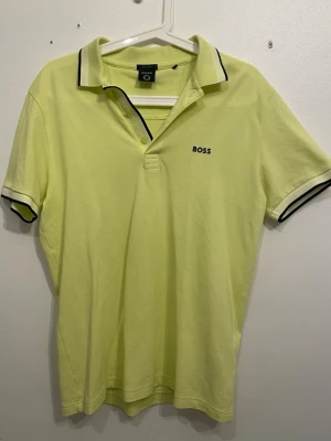 Gul pikétröja från BOSS - Fräsch gul pikétröja från BOSS med svart logga på bröstet. Tröjan har klassisk krage med svarta detaljer och korta ärmar med matchande svarta ränder. Regular fit för en avslappnad look. Perfekt till sommaren!