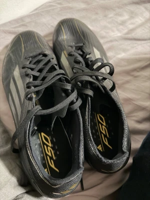 Adidas F50 svarta fotbollsskor - Säljer ett par svarta Adidas F50 fotbollsskor med klassiska vita ränder och gula detaljer. Skorna har snörning och fasta dobbar för spel på gräs. Ovandelen är i syntetmaterial och designen är smal och sportig, perfekt för snabba spelare som vill ha bra grepp och känsla på planen.