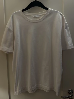 Vit basic t-shirt från Zara - En enkel och stilren vit t-shirt från Zara med rund hals och korta ärmar. Tillverkad i mjuk bomull för en skön känsla och avslappnad passform. Perfekt till jeans eller shorts för en clean look.