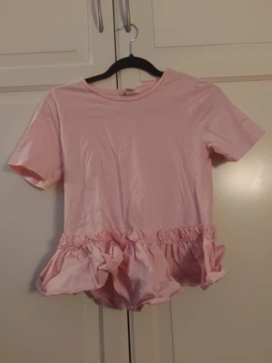 Ljusrosa topp med volangkant - Supersöt ljusrosa topp med korta ärmar och rund hals. Toppen har en bred volangkant nedtill som ger en lekfull och trendig look. Perfekt att styla med jeans eller kjol för en söt och avslappnad outfit.