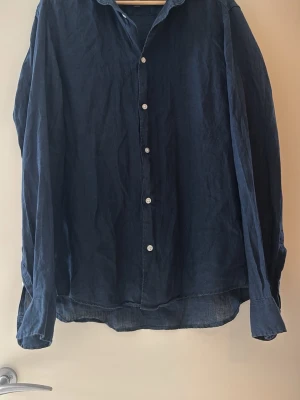 Mörkblå linneskjorta från H&M - Snygg mörkblå skjorta från H&M i linnematerial. Skjortan har slim fit-passform, klassisk krage, långa ärmar och vita knappar framtill. Perfekt för en stilren och avslappnad look.