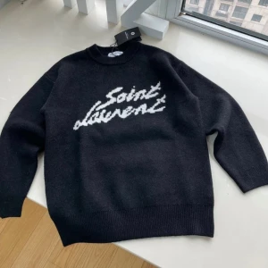 Svart stickad Saint Laurent tröja - Säljer en svart stickad tröja från Saint Laurent med vit logotyp i grafisk stil på bröstet. Tröjan har rund halsringning, ribbade muddar och är tillverkad i ett mjukt material som passar perfekt för kyligare dagar.