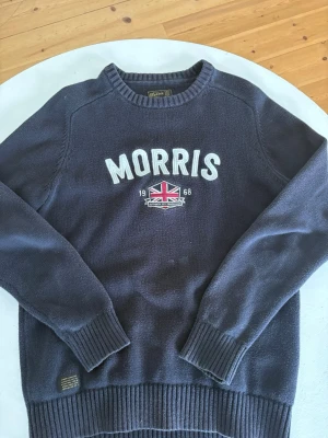 Mörkblå stickad tröja Morris - Mörkblå stickad tröja från Morris med vit broderad logga och brittisk flagga på bröstet. Tröjan har rund hals, långa ärmar och ribbade muddar vid ärmslut och nederkant. Perfekt för dig som gillar klassisk och stilren design.