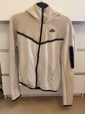 Beige Nike hoodie med dragkedja - Snygg beige hoodie från Nike med svart dragkedja och svarta detaljer. Har huva, fickor på sidorna och en extra ficka med dragkedja på ärmen. Nike-logga på bröstet och sportig design med svarta linjer över bröstet.