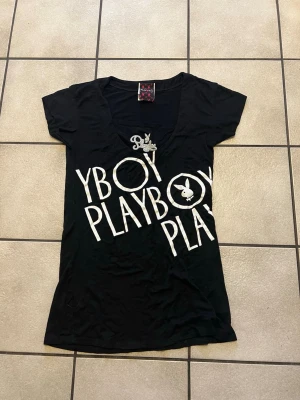 Svart klänning från Playboy storlek XS/S - Svart t-shirt från Playboy med stort vitt tryck över hela framsidan och klassisk Playboy-kanin. T-shirten har v-ringning och korta ärmar. Perfekt för dig som gillar streetstyle och vill sticka ut med en ikonisk logga.