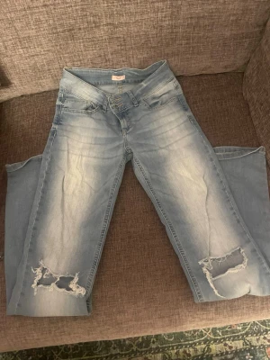 Ljusblå slitna jeans med hål - Säljer ett par ljusblå jeans med slitningar och stora hål vid knäna. Jeansen har fem fickor, knappgylf och raka ben. Materialet är klassisk denim i bomull och de har en trendig, avslappnad look.