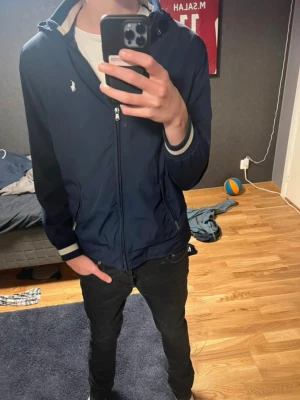 Marinblå vindjacka från Polo Ralph Lauren - Snygg marinblå vindjacka från Polo Ralph Lauren med dragkedja framtill, två sidofickor och vit broderad logga på bröstet. Jackan har ribbade muddar med vita ränder och en klassisk huva som går att ta av. Säljer pga att den inte passar längre. Hör av er vid frågor😊