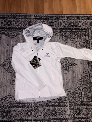 Vit Gore-Tex regnjacka Arc'teryx - Snygg vit regnjacka från Arc'teryx med huva och dragkedja framtill. Tillverkad i slitstarkt Gore-Tex-material som står emot regn och vind. Jackan har två sidofickor med dragkedja och justerbar huva för extra skydd. Perfekt för blöta dagar.