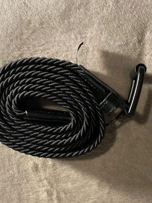 Svart flätat bälte i syntet - Snyggt svart flätat bälte med metallspänne och detaljer i konstläder. Bältet har en stilren look med twistad och vävd design som passar till många outfits. Perfekt accessoar för att lyfta din stil. Jättebra bälte perfekt går bra att använda med alla sorts outfit. den funkar perfekt för både killar och tjejer