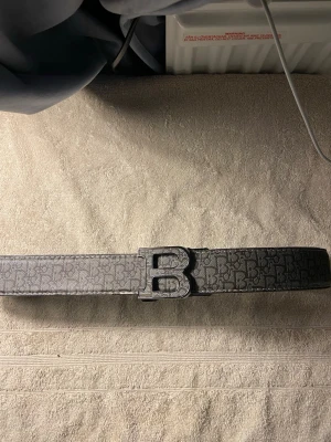 Svart Balmain bälte med B-spänne - Snyggt svart bälte från Balmain med ett stort B-format metallspänne. Bältet har ett diskret mönster med Balmain-loggor över hela remmen och är gjort i skinn. Perfekt accessoar för att lyfta vilken outfit som helst.