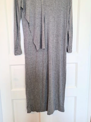 Grå Dagmar-klänning i ullmix - Stilren, mjuk & högkvalitativ - Strl S - Stilren grå klänning från Dagmar i mjuk modal/ull-mix. V-ringning och asymmetrisk drapering som ger ett fint fall. Perfekt både till vardag och jobb. Storlek S.  Mått: Bystomkrets ca 96 cm och längd från axel 118 cm Skick: Jättefin fortfarande och inte kunnat hitta några fel på den. Finns några enstaka noppror. Väldigt finstickat material så jag förbehåller mig att jag kanske missat en tråddragning eller dylikt. 