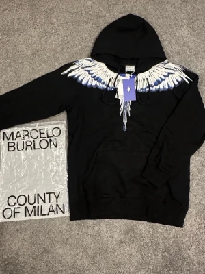 Svart hoodie Marcelo Burlon med vingar - HELT NY!!Svart hoodie från Marcelo Burlon County of Milan med ikoniskt vingtryck i vitt och blått över axlar och bröst. Klassisk huva med dragsko och känguruficka framtill. Stilsäker och unik design som sticker ut i mängden.
