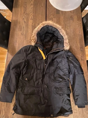 Svart vinterjacka från Parajumpers - Säljer en svart vinterjacka från Parajumpers med avtagbar huva och fuskpäls. Jackan har flera stora fickor, gul detalj vid dragkedjan och patch på bröstet. Perfekt för kalla dagar och riktigt snygg streetstil.