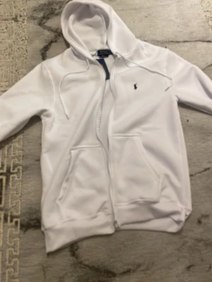 Polo Ralph Lauren Fleece Full-Zip Hoodie - Snygg vit hoodie från Polo Ralph Lauren med dragkedja framtill, två fickor och justerbar huva med snören. Tillverkad i stretchigt material för extra komfort. Klassisk liten Polo-logga broderad på bröstet. Perfekt för en clean och stilren look.