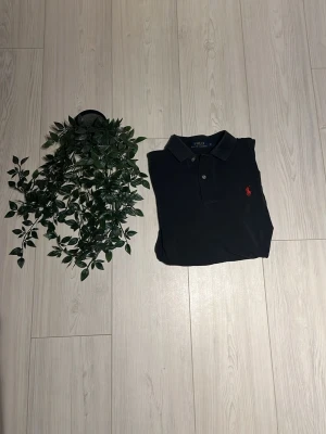 Svart pikétröja från Polo Ralph Lauren - Klassisk svart pikétröja från Polo Ralph Lauren med krage och två knappar framtill. Tröjan har den ikoniska röda logotypen broderad på bröstet. Perfekt till jeans eller shorts för en clean och stilren look.