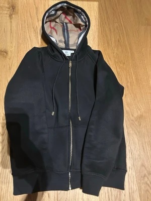 Svart hoodie med dragkedja från Burberry - Snygg svart hoodie från Burberry med dragkedja framtill och klassiskt rutigt foder i huvan. Hoodien har justerbara snören och fickor på magen. Perfekt för dig som vill ha en stilren och exklusiv look med en touch av brittisk design.