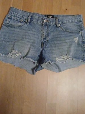 Blå denimshorts med slitningar 157 - Säljer ett par blå denimshorts från 157 med råa kanter och slitna detaljer framtill. Klassisk femficksmodell med knapp och dragkedja. Perfekta för sommaren och ger en avslappnad look.