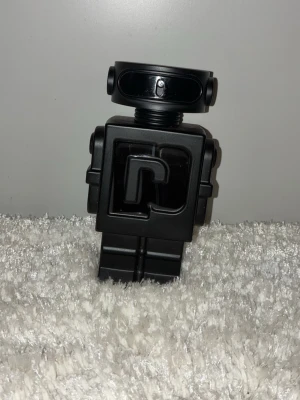 Paco Rabanne Phantom parfym robotflaska 90ml - Cool parfymflaska från Paco Rabanne i form av en svart robot. Flaskan har en futuristisk design med tydliga detaljer och ett stort 'P' på bröstet. Tillverkad i plast och metall-look, perfekt för dig som gillar unika och snygga parfymflaskor.