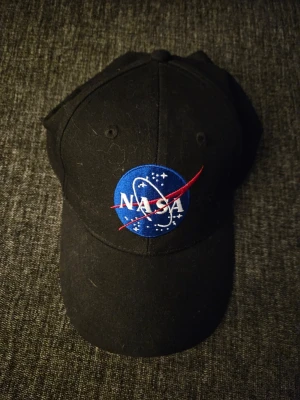 Svart NASA keps med broderad logga - Snygg svart keps med den klassiska NASA-loggan broderad framtill i blått, vitt och rött. Kepsen har böjd skärm och justerbar passform baktill. Perfekt för dig som gillar rymden och vill ha en cool accessoar.