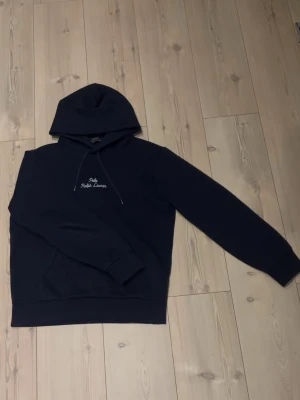 Mörkblå hoodie från Polo Ralph Lauren - Mörkblå hoodie från Polo Ralph Lauren med broderad vit logga på bröstet. Klassisk modell med huva och dragsnören samt ribbade muddar vid ärmslut och midja. Perfekt för en avslappnad och stilren look.
