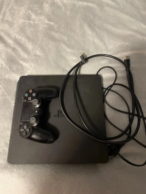 PlayStation 4 Slim med handkontroll - Svart PlayStation 4 Slim med en matchande svart DualShock 4-handkontroll och HDMI-kabel. Konsolen har en stilren, fyrkantig design i matt plast och är perfekt för gaming och streaming. Allt du behöver för att komma igång med spelandet direkt.